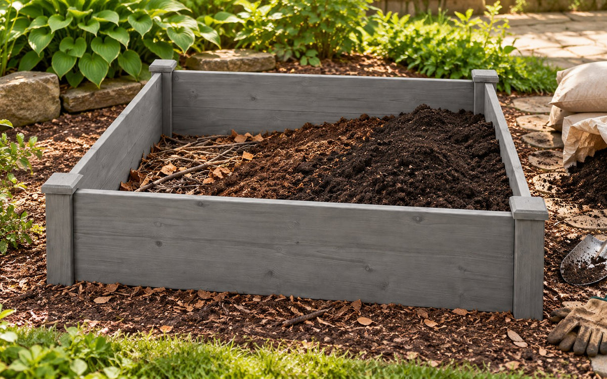 A low gray wooden raised bed is being filled with soil in a backyard garden.
